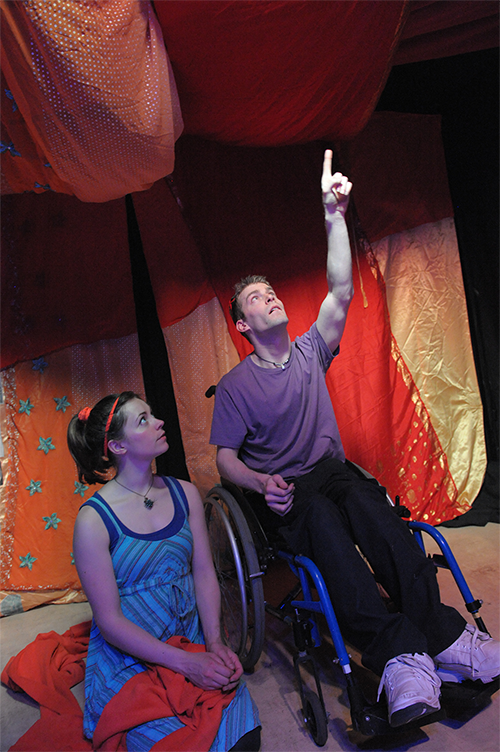 2 people looking up on stage, and one person pointing upwards. There are orange, gold, and red fabrics in the background and hanging from the ceiling.